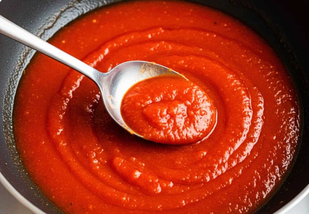 A metal spoon stirring thick tomato sauce in a black frying pan, showing the smooth and vibrant texture.