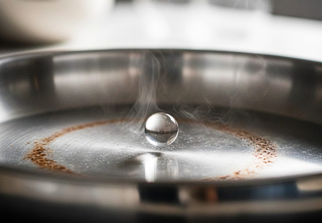 A macro shot of a single water droplet perfectly spherical and hovering over a hot stainless steel pan.