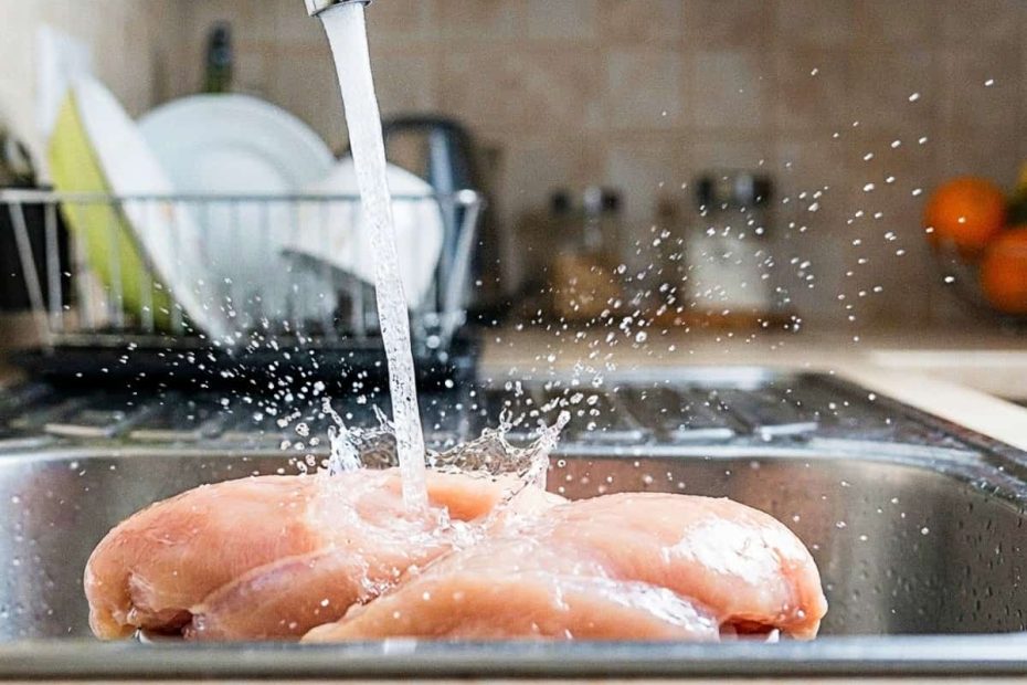 Close-up of raw chicken being washed under a faucet showing water splashes in a kitchen sink