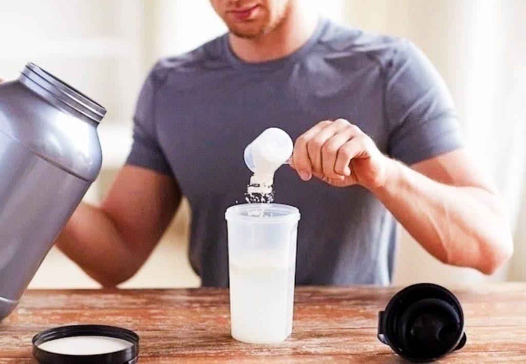 A candid action shot under natural light of a fit man in a grey t-shirt holding a massive grey supplement tub. His right hand holds a scoop over a clear shaker bottle filled with dense milk, creating a white cloud of powder verted. The shaker sits on a rustic wooden table, with the black lid off next to it. Focus is sharp on the powder stream. No whole foods are present, only technical gym preparation.