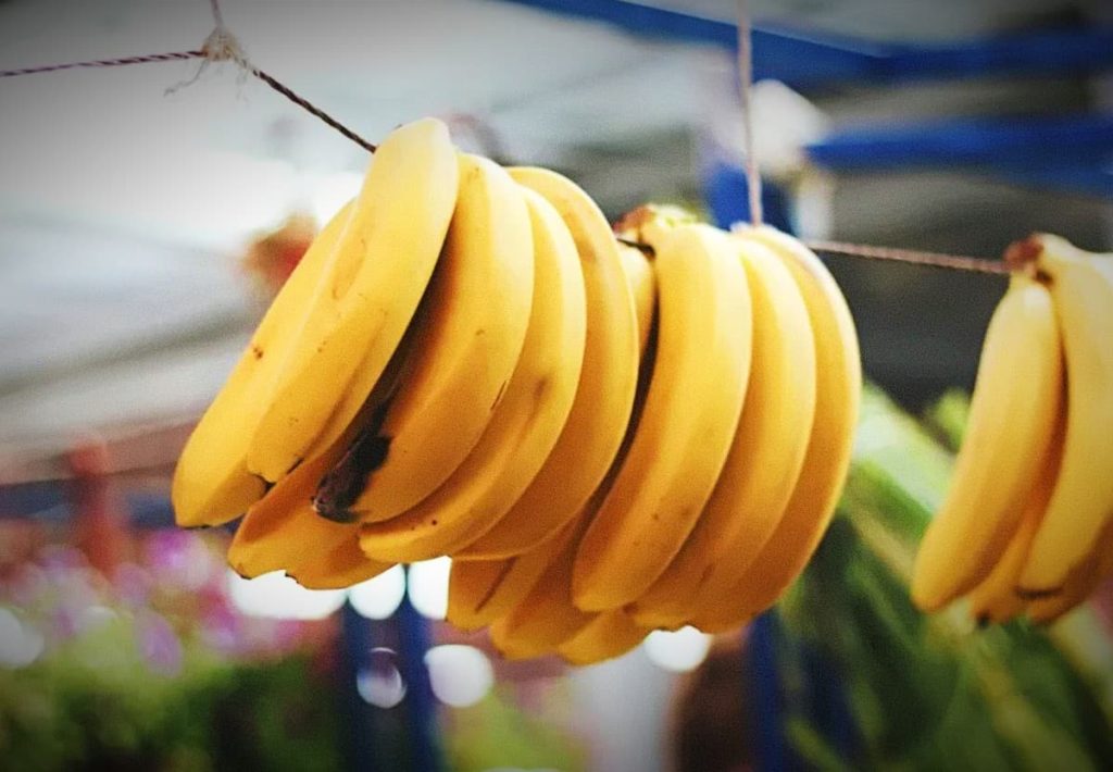 Several bunches of yellow bananas hanging from strings at an outdoor market with a blurred background.
