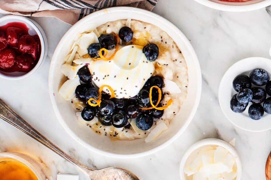 A top-down view of a white ceramic bowl filled with creamy oatmeal, topped with fresh blueberries, orange zest, coconut flakes, and a dollop of Greek yogurt.