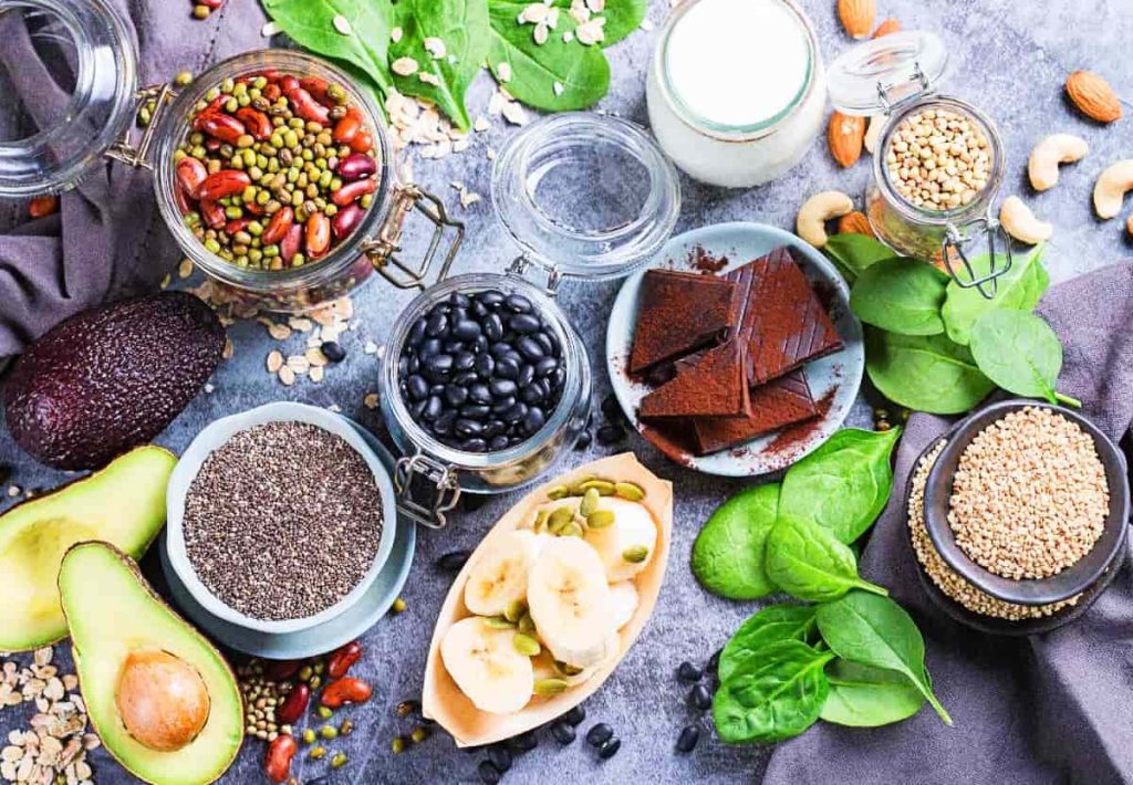 A top-down spread of magnesium-rich foods including dark chocolate, black beans, pumpkin seeds over bananas, avocado, spinach, and hemp seeds.