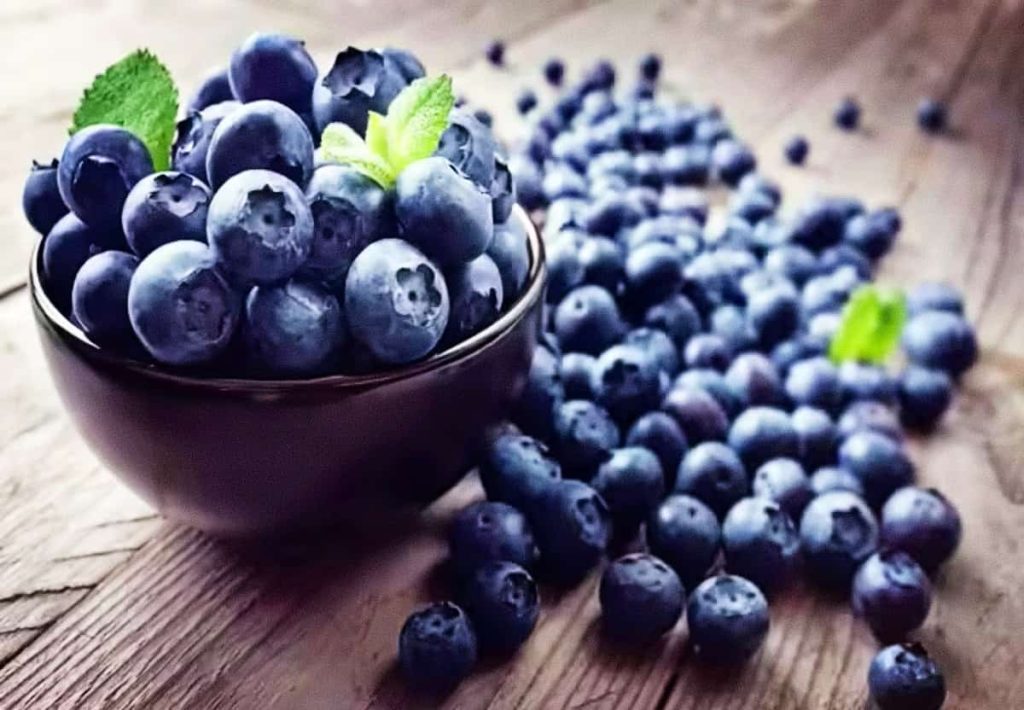 A dark bowl overflowing with fresh blueberries on a rustic wooden table, with many berries scattered around, garnished with mint leaves.