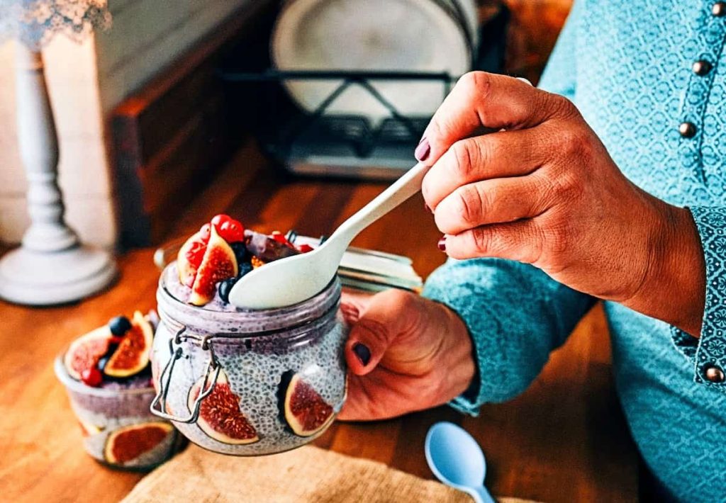A close-up of a person in a teal shirt holding a glass jar of chia seed pudding topped with fresh figs and berries, illustrating soluble fiber's role in GLP-1 release.