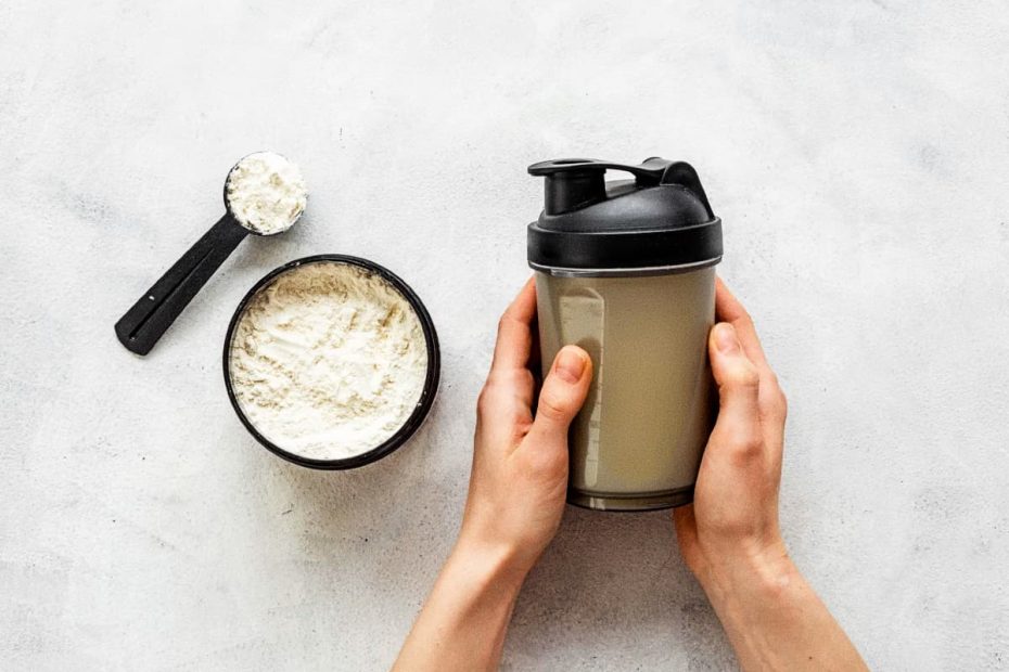 Top-down view of a person's hands holding a protein shaker next to an open container of white whey protein powder and a measuring scoop on a white stone background.