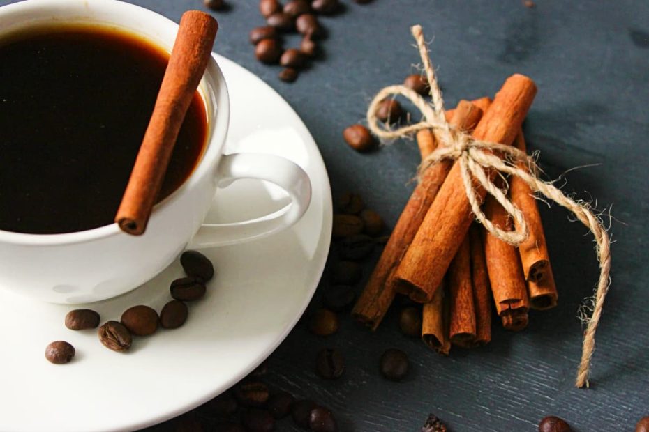 A white cup of black coffee with a cinnamon stick inside, next to a bundle of cinnamon sticks and scattered coffee beans on a dark surface.