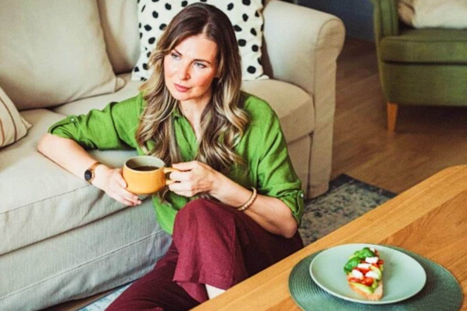 A woman in a green shirt and burgundy pants sitting on the floor with a mug of coffee and a piece of avocado toast with mozzarella, representing healthy metabolic habits.