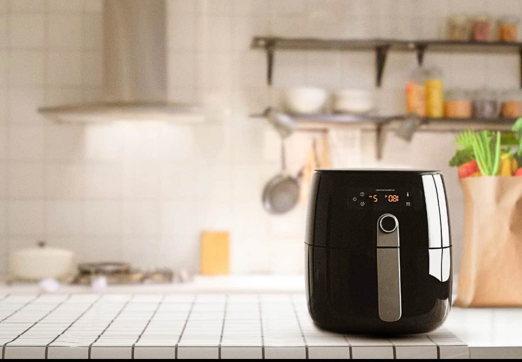 A modern black air fryer sitting on a checkered tablecloth in a bright, well-organized kitchen with a bag of vegetables in the background.