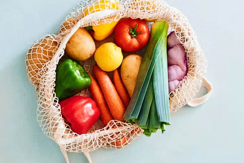 A top-down view of a white cotton mesh produce bag filled with colorful vegetables like bell peppers, carrots, tomatoes, and leeks on a light blue background.