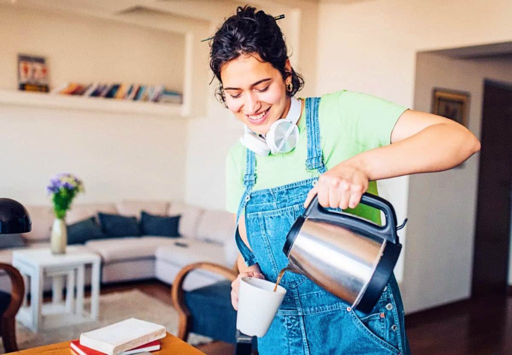 A young woman in denim overalls and a green t-shirt with headphones around her neck smiling while pouring coffee into a mug from a silver electric kettle.