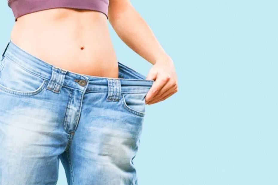 A woman in a purple sports bra holding out the waistband of her blue jeans to show how much weight she has lost around her waist.
