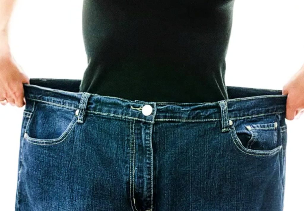 Close-up of a person in a black shirt pulling on the sides of oversized denim jeans to demonstrate significant fat loss and space in the garment.