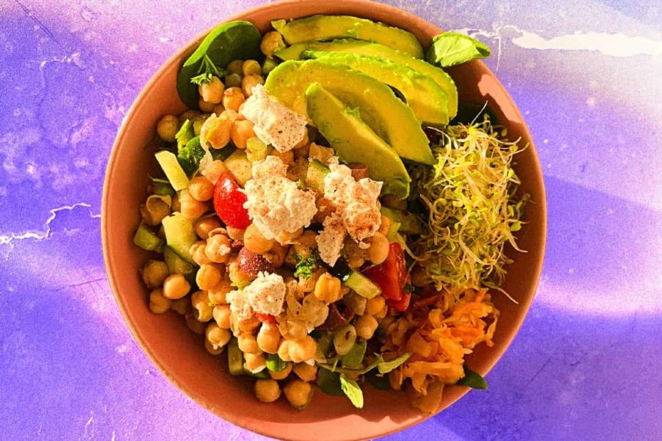 A vibrant high-protein bowl featuring chickpeas, sliced avocado, fresh sprouts, cherry tomatoes, and crumbled feta cheese on a purple background.