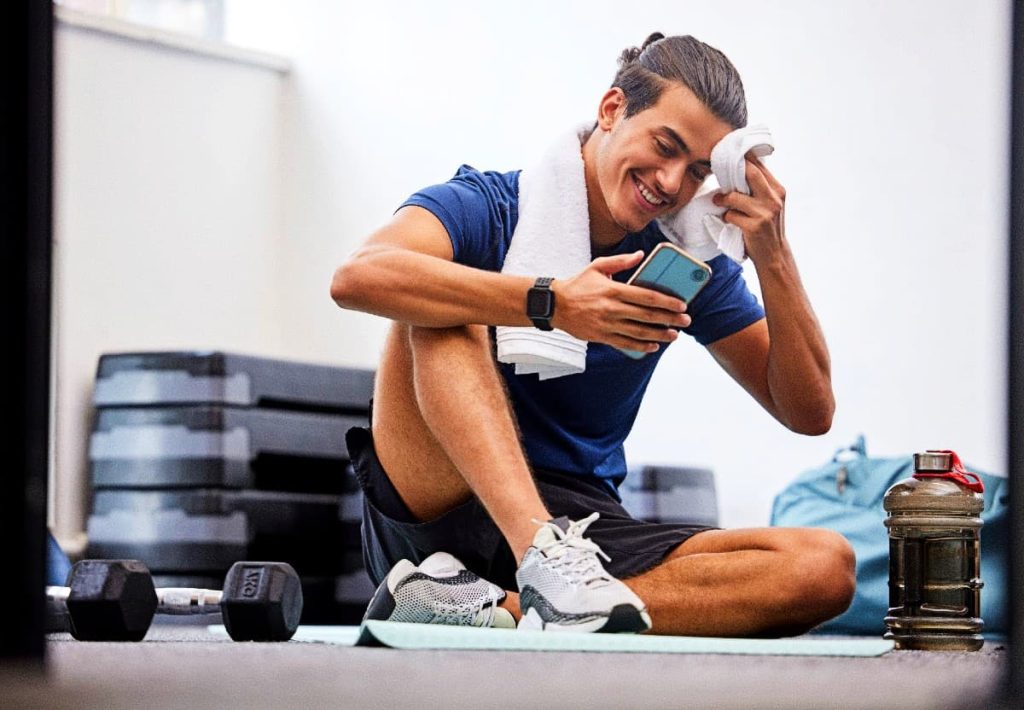 A young man sitting on a gym floor after a workout, smiling while checking his phone and wiping sweat with a towel, with dumbbells and a water bottle nearby.