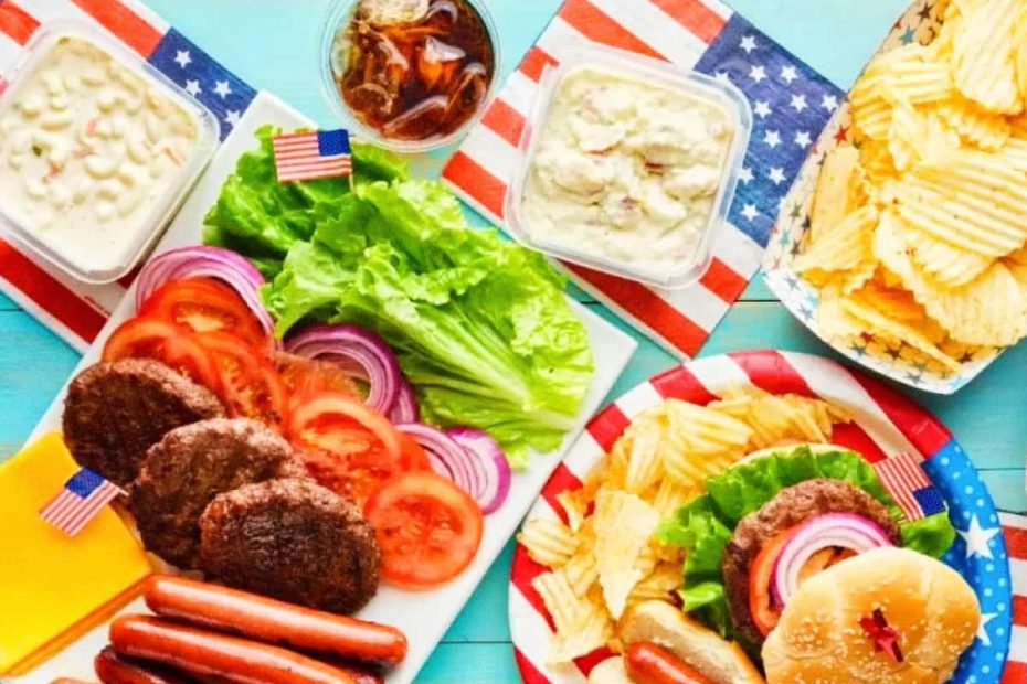 A colorful top-down spread of burgers, sausages, french fries, and potato chips with American flag picks on a blue background.