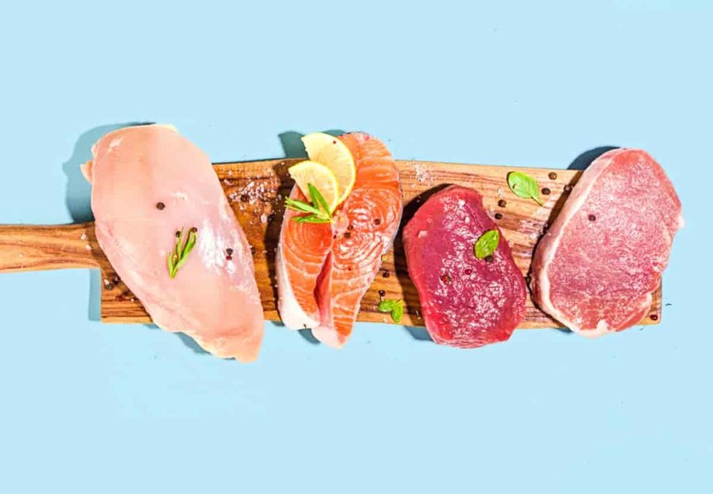 A top-down view of chicken breast, salmon steak with lemon slices, a beef medallion, and a pork chop arranged on a wooden cutting board against a light blue background.