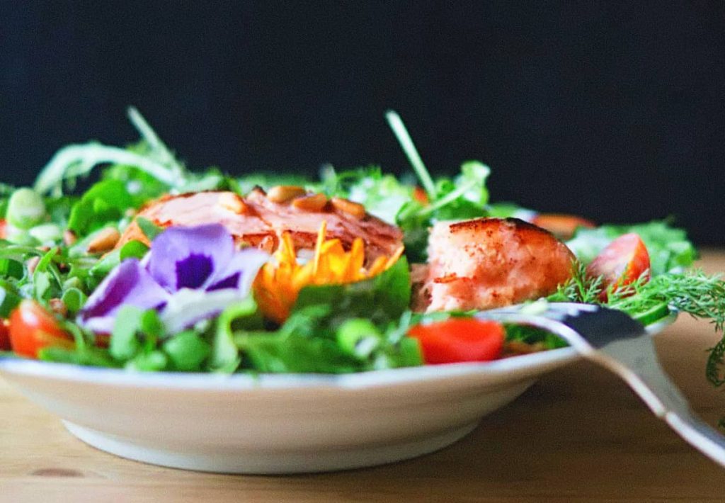 A gourmet salad featuring grilled salmon, fresh greens, cherry tomatoes, and edible purple flowers on a white plate.