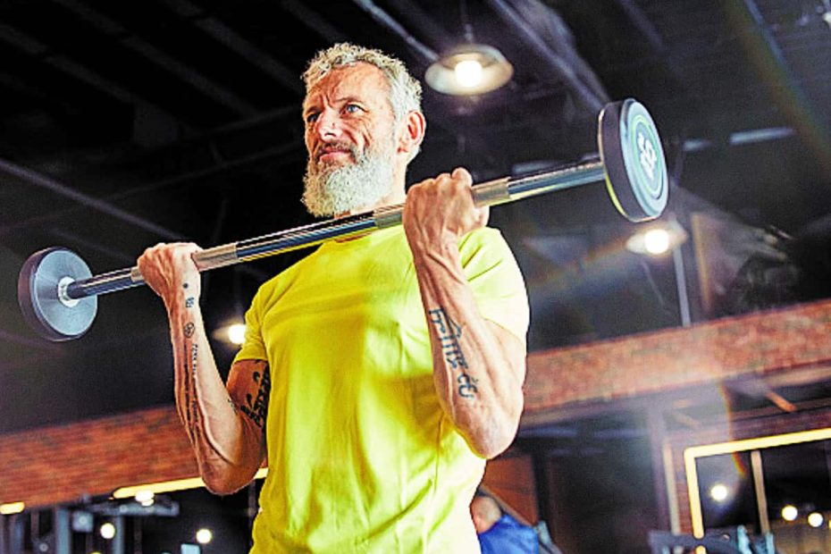 A middle-aged man with gray hair and a beard, wearing a bright yellow t-shirt, performing a barbell curl with focused determination in a modern gym with warm lighting.