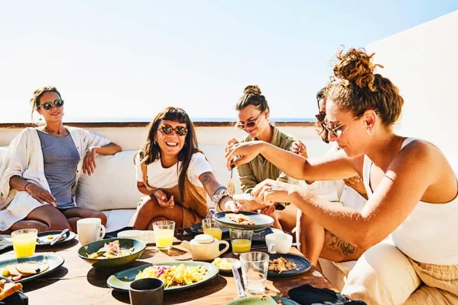 A group of five women laughing and sharing a healthy outdoor brunch on a sunny terrace, symbolizing the natural satiety and social aspect of the GLP-1 lifestyle.
