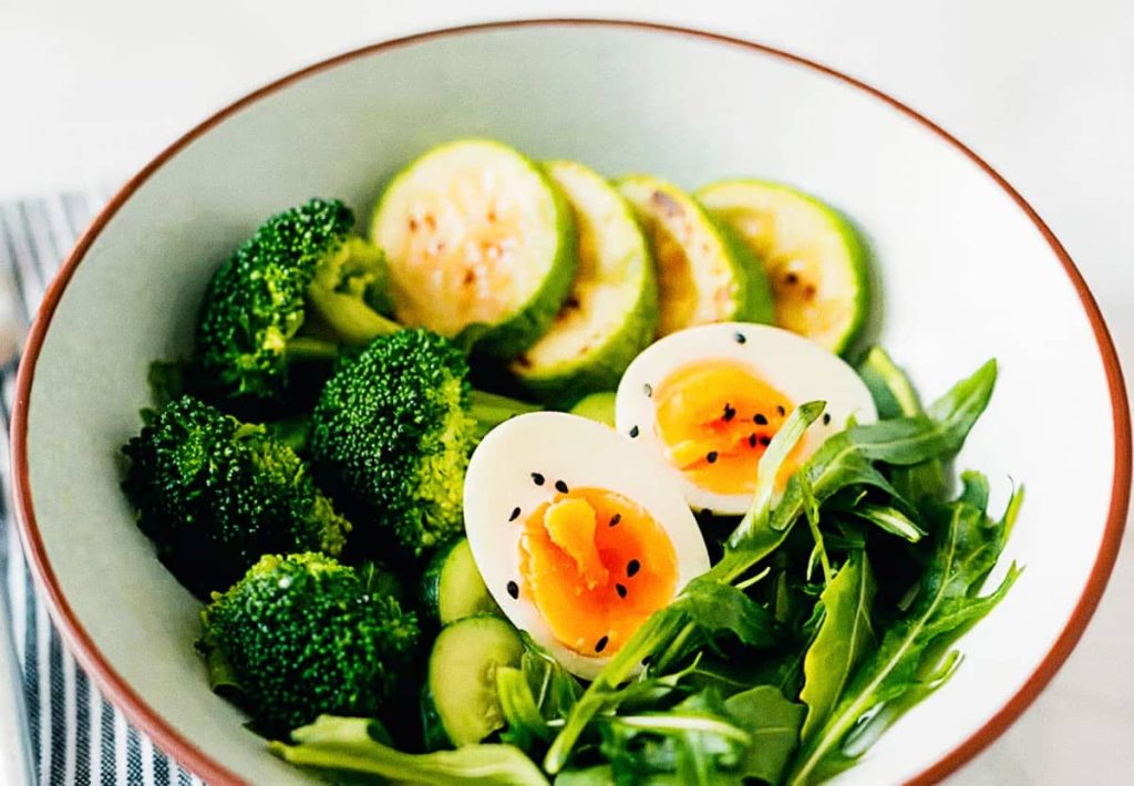 A white bowl filled with soft-boiled eggs, grilled zucchini, fresh broccoli, and arugula, demonstrating a high-protein, nutrient-dense meal.
