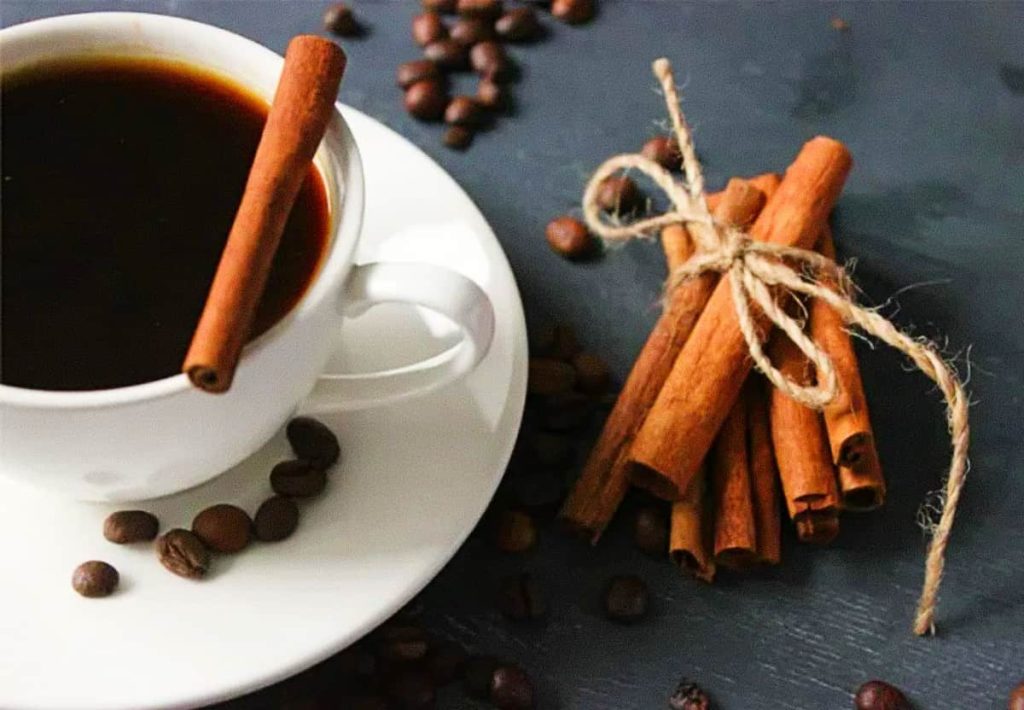 A white cup of black coffee on a saucer, with a whole cinnamon stick resting on the rim and a bundle of cinnamon sticks tied with twine in the background.