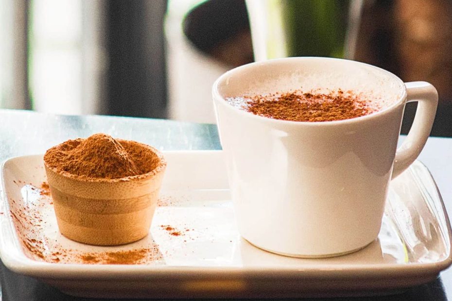 A white cup of coffee topped with a thick layer of ground cinnamon, placed on a white plate next to a small wooden bowl filled with extra cinnamon powder.