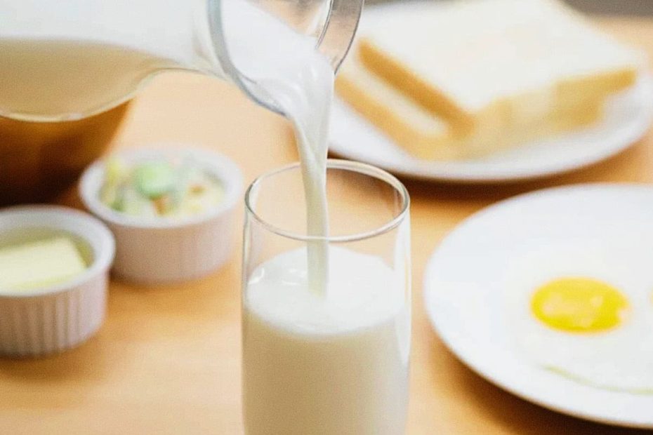 Milk being poured from a glass jug into a tall clear glass on a wooden table, with butter and bread in the soft-focus background.