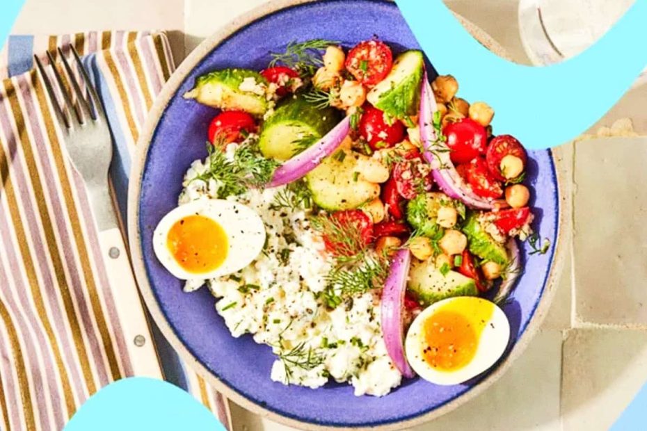 A blue bowl filled with cottage cheese, sliced cucumbers, cherry tomatoes, chickpeas, red onion, and two soft-boiled egg halves.