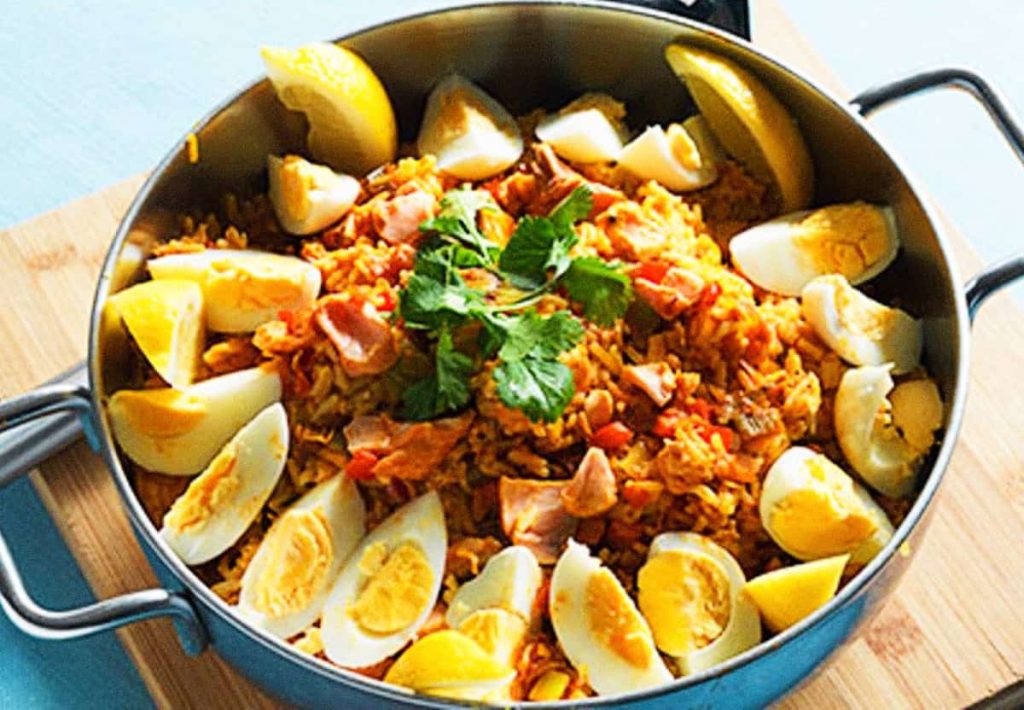 A large metal pan containing a savory mixture of chickpeas, rice, and vegetables, topped with hard-boiled egg wedges and lemon slices.