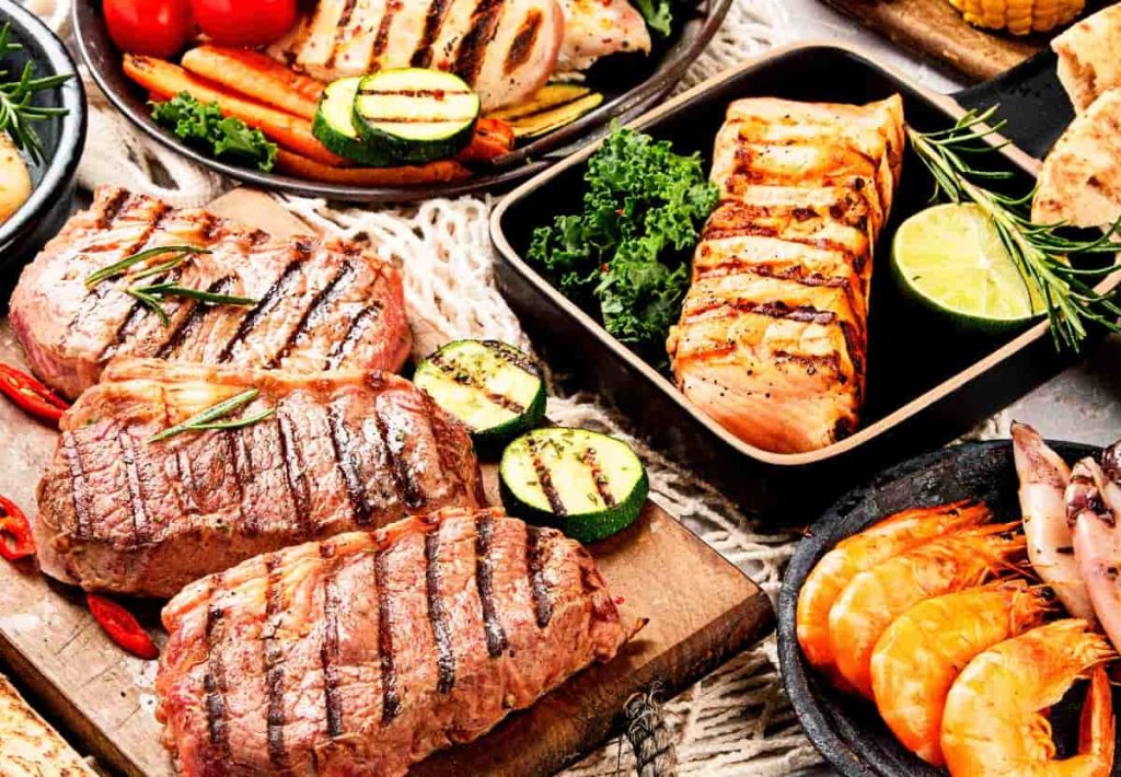 Grilled lean steaks served on a wooden board alongside grilled zucchini, tomatoes, and other colorful vegetables.
