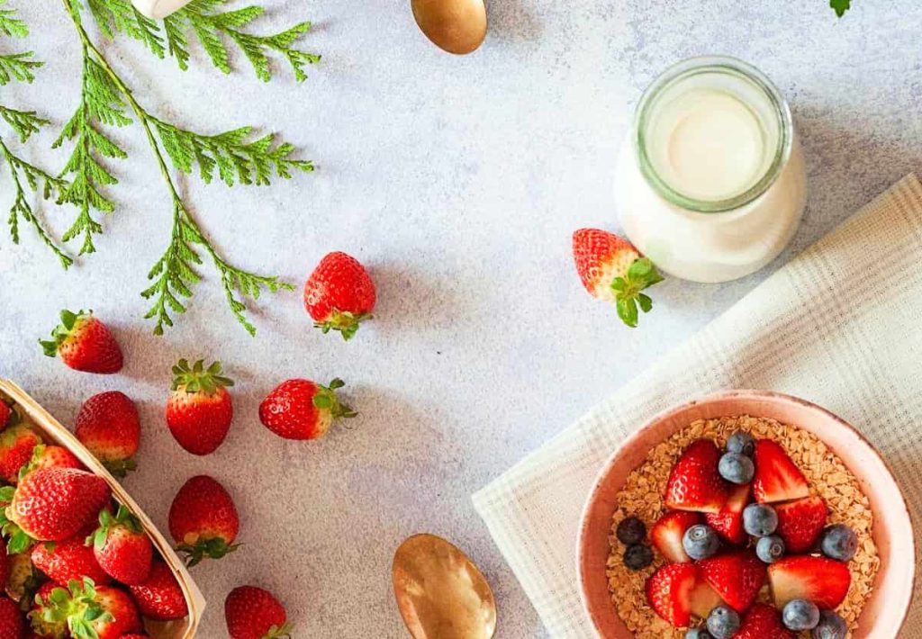 Top-down view of a healthy breakfast bowl with strawberries, blueberries, and oats next to a small bottle of fresh milk and green sprigs.