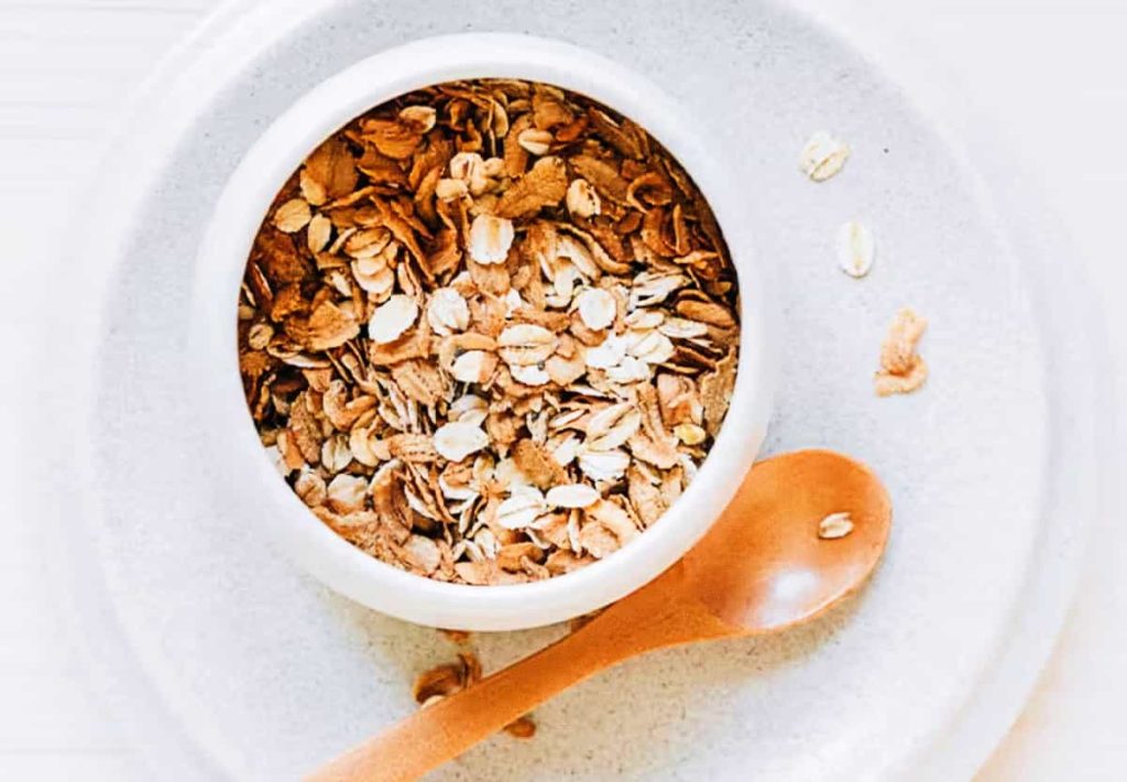 Top-down view of a small white ceramic bowl containing dry rolled oats on a light-colored plate with a wooden spoon.