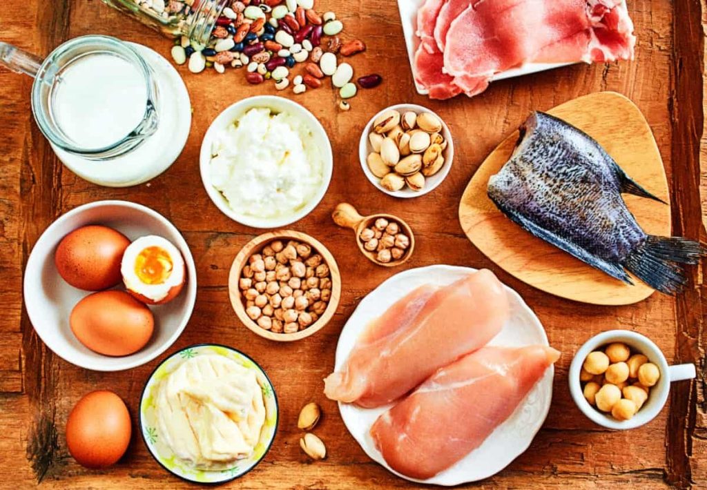 A top-down view of a white baking dish containing salmon steaks, roasted tomatoes, brussels sprouts, and broccoli with lemon wedges on a rustic white wood surface.