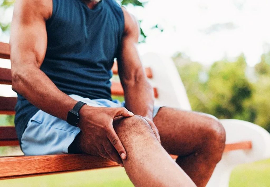 A close-up shot of an athletic man in a dark tank top sitting on a wooden park bench, with his hand firmly holding his knee, suggesting post-workout pain or joint inflammation.
