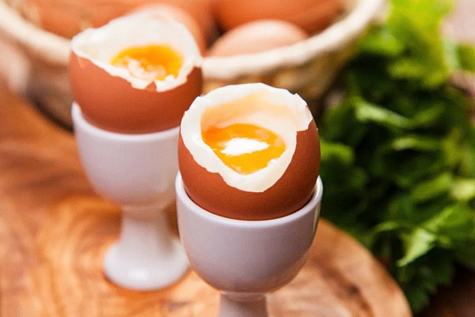 Two soft-boiled eggs in white egg cups with golden runny yolks, served on a wooden board with fresh herbs in the background.