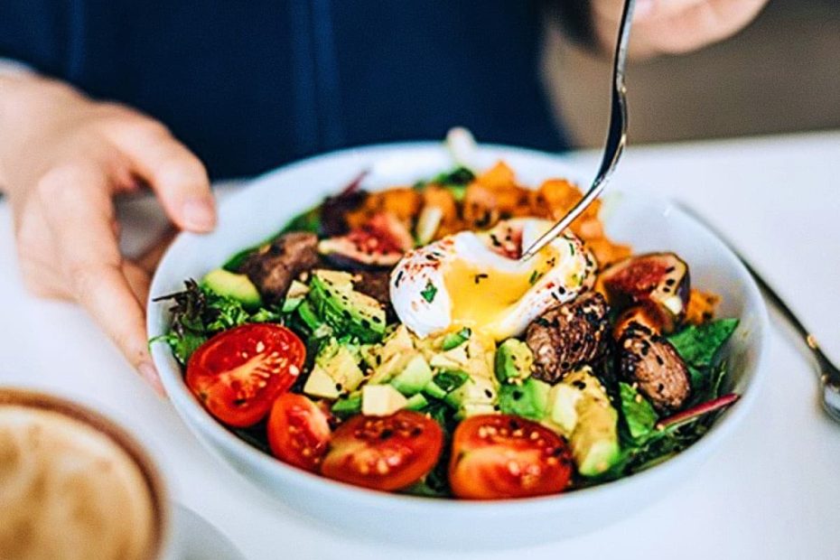 A white bowl filled with a diverse salad featuring a poached egg with a runny yolk, avocado slices, cherry tomatoes, and seared protein pieces.
