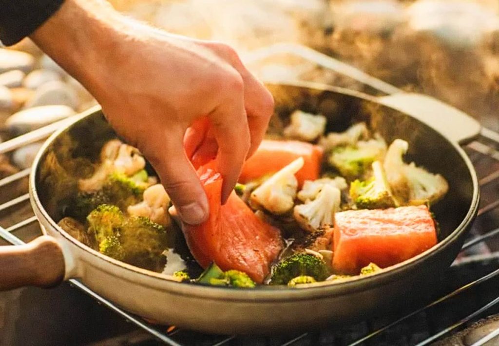 A person’s hand placing a piece of raw salmon into a frying pan already filled with broccoli and cauliflower florets.