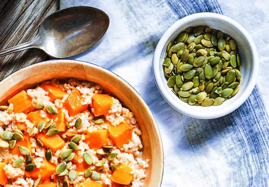 A top-down view of a bowl of cooked grains topped with diced pumpkin and seeds, next to a small white bowl filled with raw pumpkin seeds.