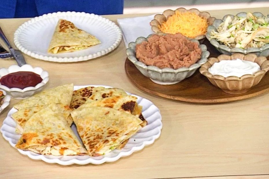 A wooden table with several plates containing folded crepiocas, bowls of mashed beans, shredded chicken, grated cheese, and a white dipping sauce.