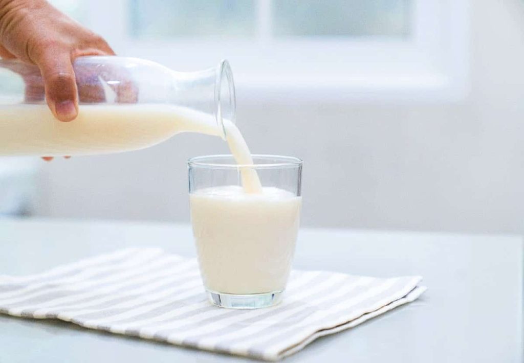 A close-up shot of a hand pouring fresh white milk from a glass bottle into a clear glass sitting on a striped napkin.
