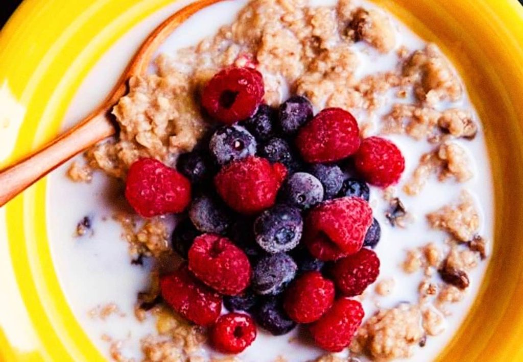 A yellow bowl of oatmeal with a wooden spoon, showing a pool of milk around the edges and topped with raspberries and blueberries.