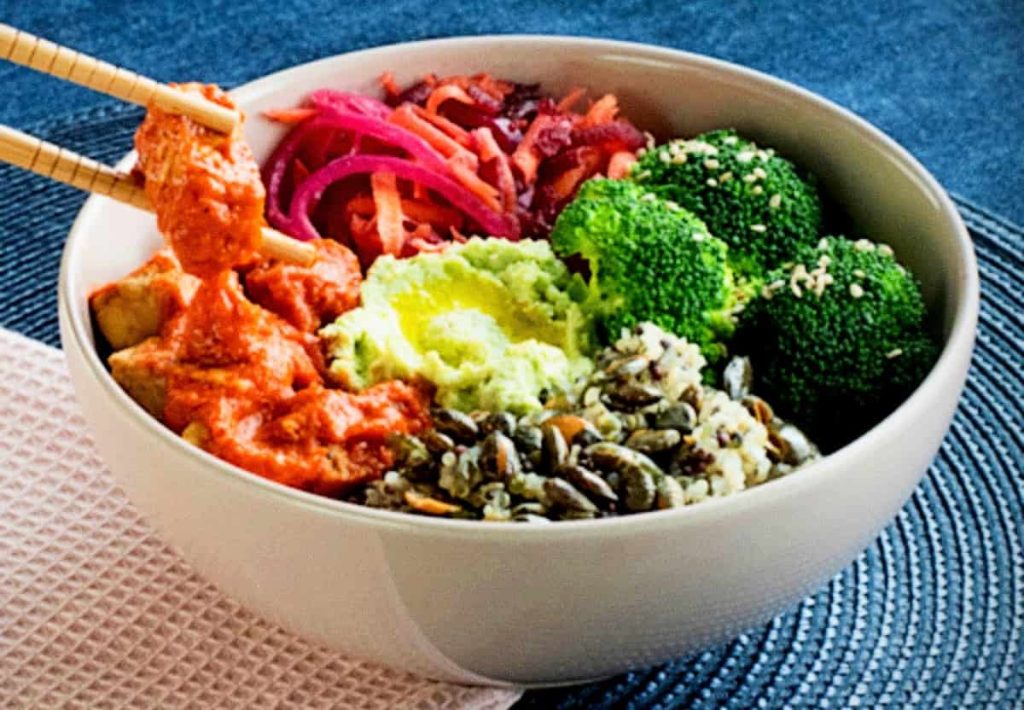 A close-up of a person using chopsticks to pick up a piece of saucy tofu from a bowl with steamed broccoli, pickled red onions, and pumpkin seeds.