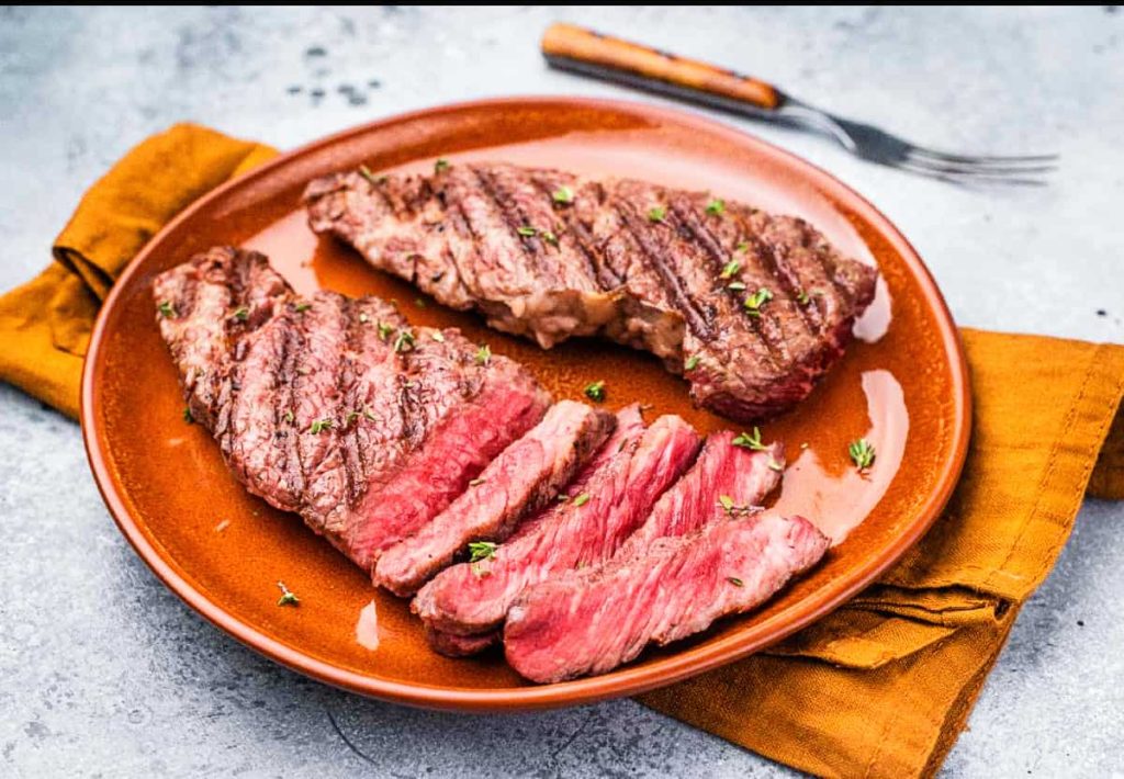 Two grilled steaks, one sliced to show a medium-rare center, served on a brown ceramic plate with a sprig of thyme and a fork in the background.