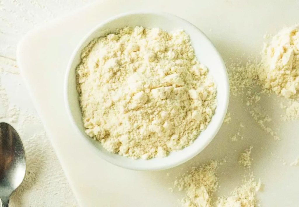 A white ceramic bowl filled with fine white protein powder on a marble surface, with a silver spoon visible on the side.