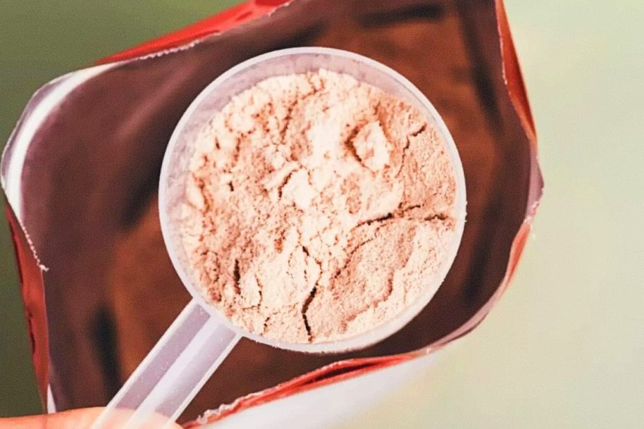 A top-down close-up shot of a plastic scoop filled with chocolate-colored protein powder inside a red supplement bag.