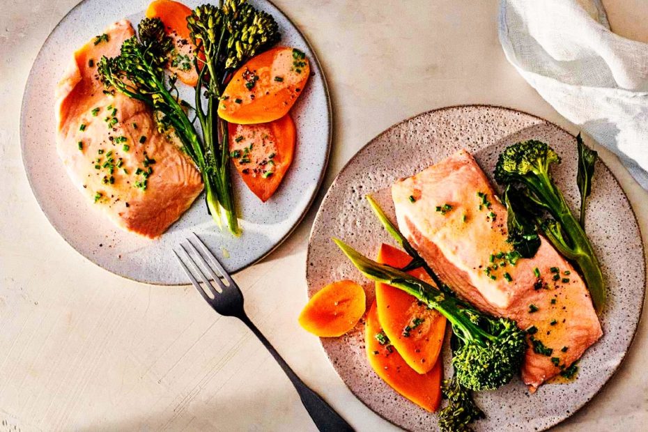 op-down shot of two rustic grey plates with seared pink salmon fillets, roasted sweet potato slices, and broccolini, garnished with green herbs. A dark fork and light linen napkin are nearby on a minimalist surface.