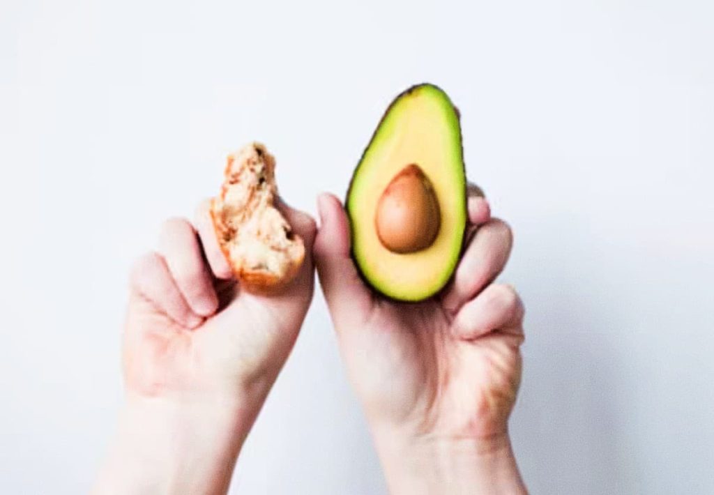 Close-up of hands holding a fresh avocado half with the seed and a piece of bread, symbolizing the integration of the pit into a healthy diet.