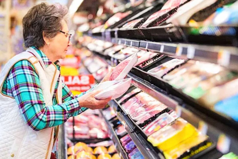 A senior woman in a supermarket aisle carefully inspecting labels on packaged red meat to identify quality and processing levels.