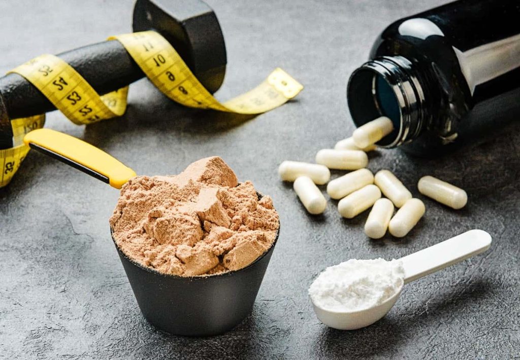A collection of fitness supplements including a scoop of brown powder, a spoon of white creatine powder, white capsules, and a dumbbell with a measuring tape.